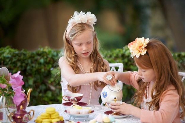 Two young girls at a tea party sit at a table adorned with a floral tablecloth. The girl on the left is pouring tea into a cup. Both wear floral headbands and pearls. The table has teacups, a teapot, yellow macarons, and a vase with pink flowers.
