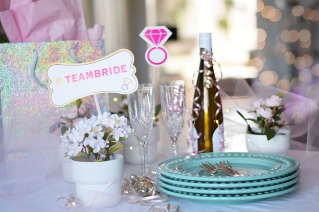 A bridal shower table setup features a stack of turquoise plates, a bottle with a ribbon, champagne glasses, small potted flowers, and a sign reading #TEAMBRIDE alongside a pink diamond ring decoration.