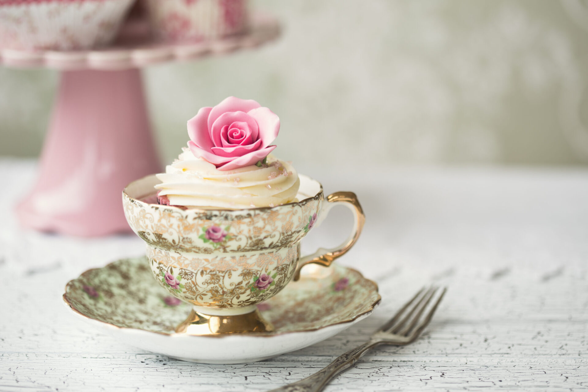 A cupcake with white frosting and a pink sugar rose sits in an ornate teacup on a matching saucer. The setup is on a textured white surface with a blurred pink cake stand in the background. A fork lies beside the saucer.