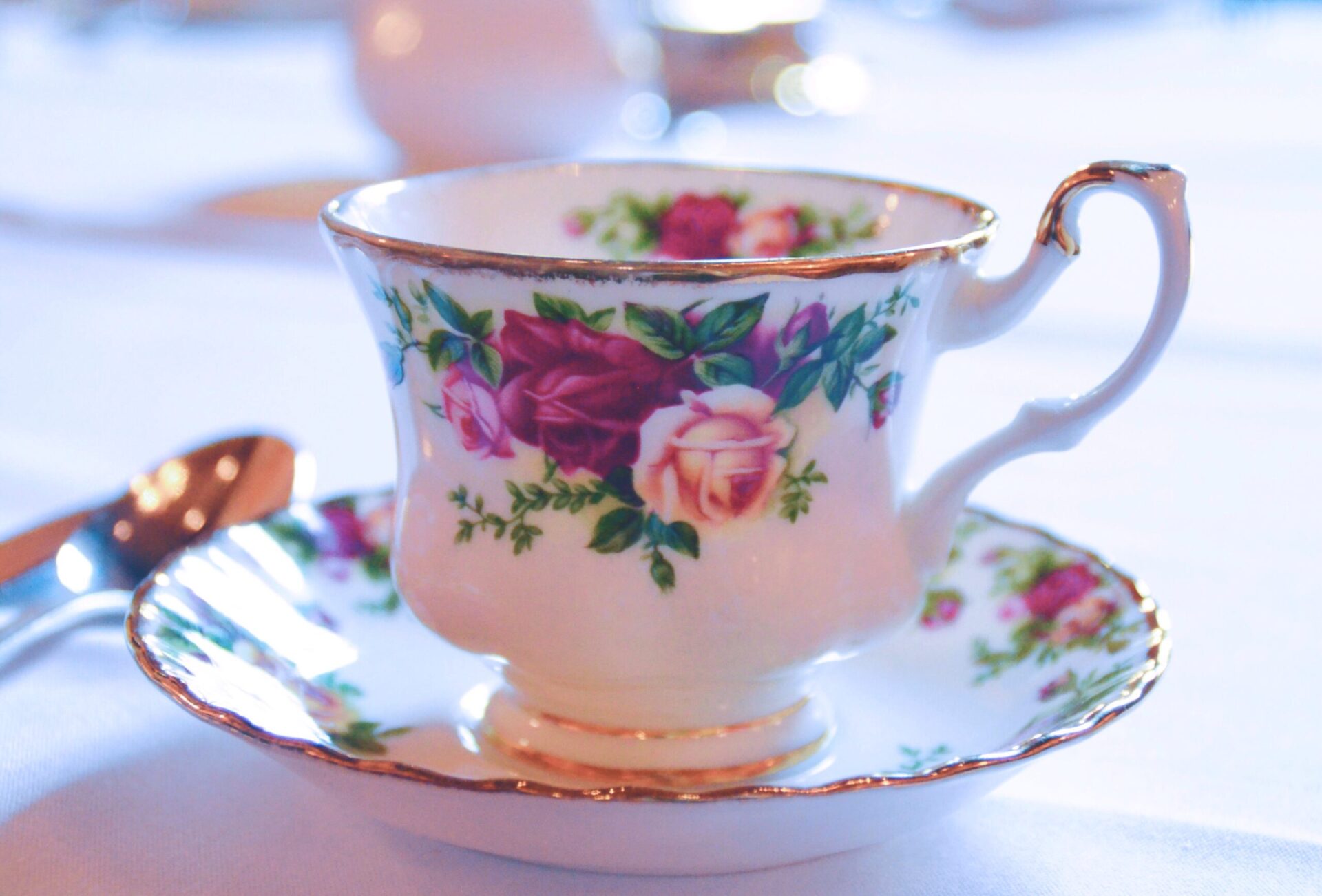 Close-up of an elegant teacup and saucer set on a white tablecloth. The teacup is decorated with a pattern of roses in shades of pink and purple, with gold accents on the rim and handle. A silver spoon rests on the table beside it.
