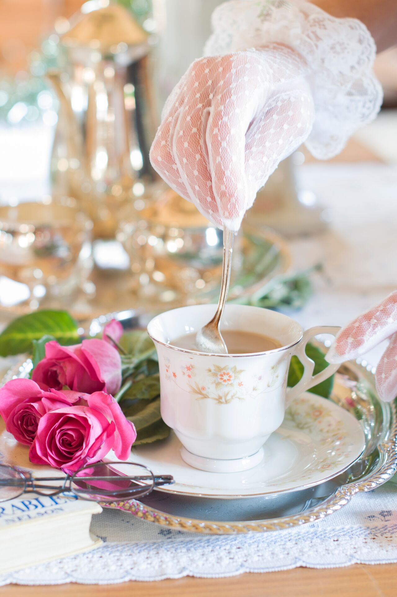 A delicate tea scene features a gloved hand stirring tea in a floral porcelain cup on a saucer. Pink roses and a book lie beside the silver tray, which has a lace tablecloth. A silver teapot is in the background.