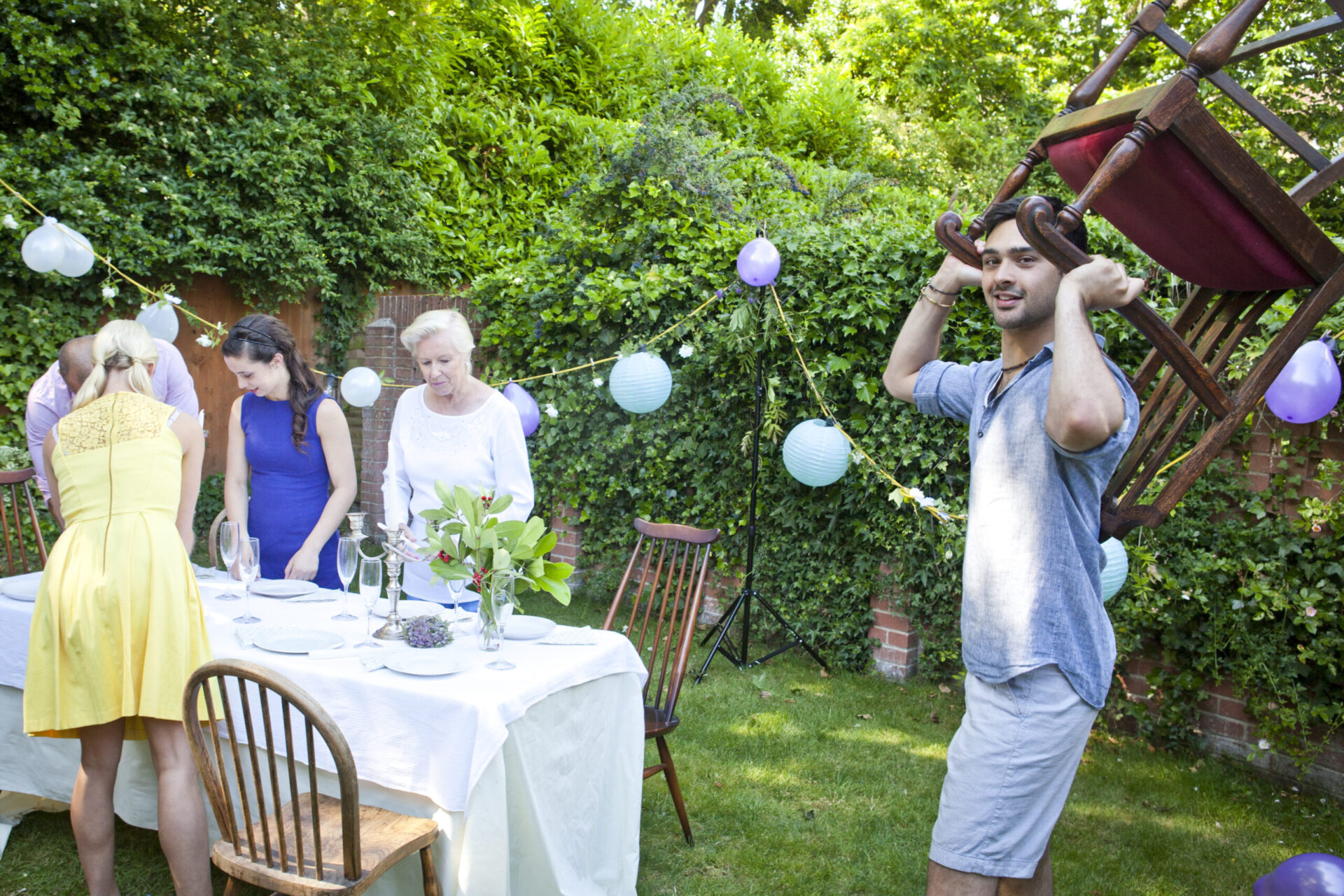 A group of people is setting up an outdoor dinner party. A man carries a chair on his shoulders, and others are arranging a table with a white tablecloth. Festive string lights and balloons decorate the garden area.
