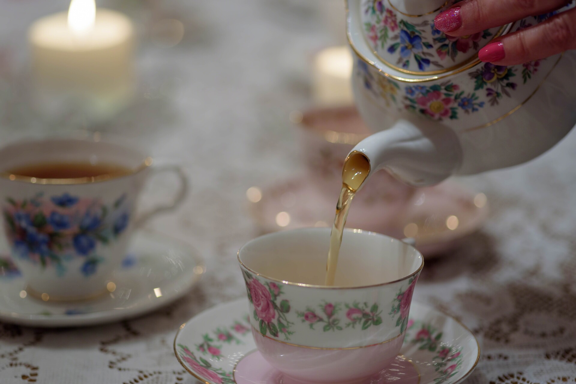 A woman pours tea from a floral-patterned teapot into a matching teacup on a lace tablecloth. Other teacups and lit candles are softly blurred in the background, creating a cozy atmosphere.