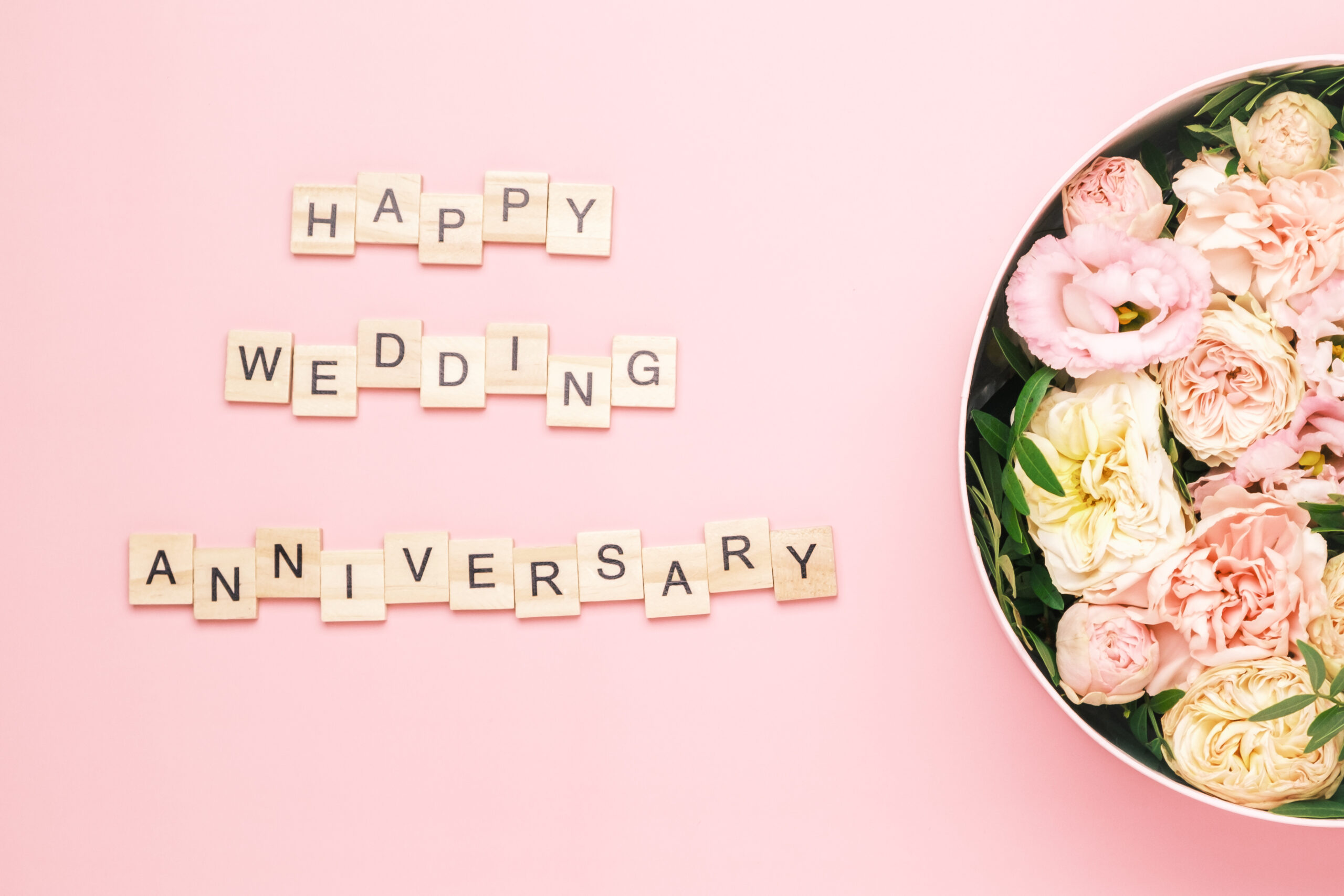 Scrabble tiles spelling “happy wedding anniversary” on a pink background, with a round box of assorted pink and white flowers to the right.