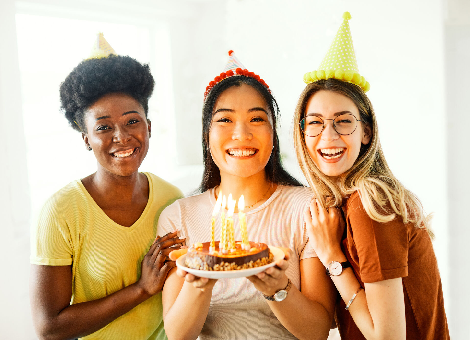 Three smiling women wearing party hats celebrate a birthday. the woman in the middle holds a cake with lit candles, while the other two stand close, all looking happy in a bright, cheerful setting.