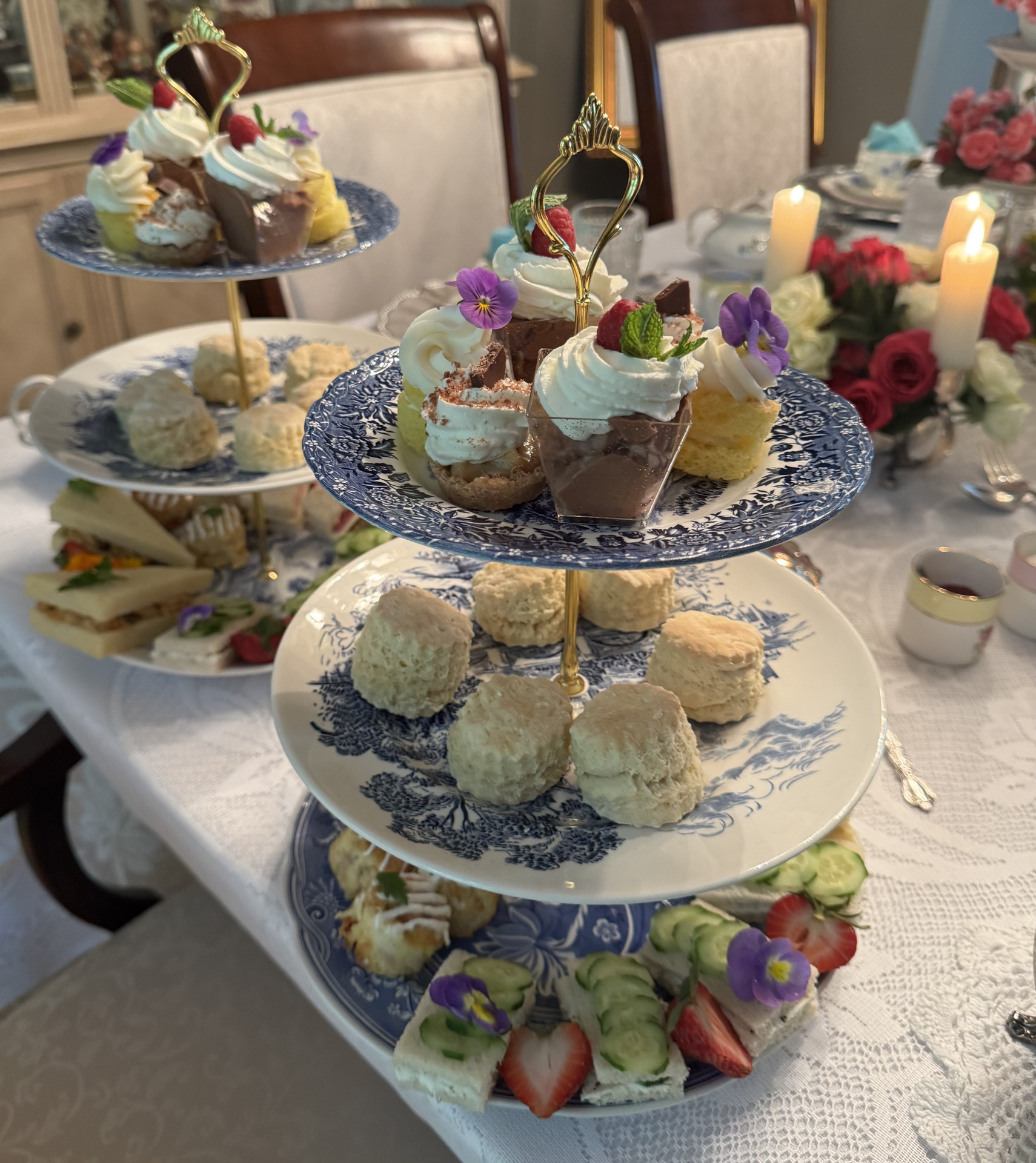 Three tiered trays with assorted pastries, scones, sandwiches, and fruit are set on a lace covered table. floral arrangements, lit candles, and teacups complete the elegant afternoon tea setting.