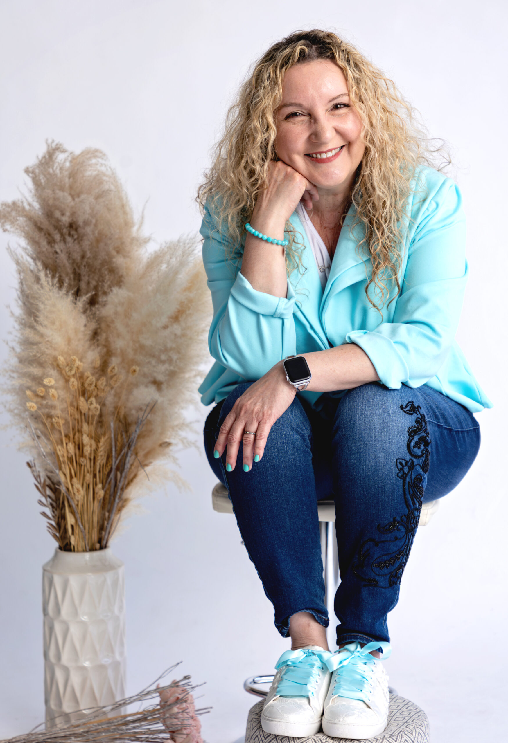 A smiling woman with curly blonde hair sits on a stool, wearing a light blue blazer, jeans with embroidery, and white sneakers. she rests her head on her hand. dried pampas grass in a vase is beside her.